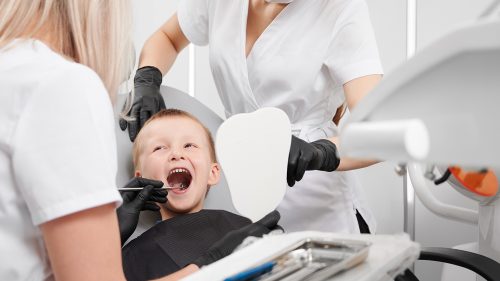 Adorable little boy sitting in dental chair while two female dentists checking kid teeth. Dentist examining little boy teeth with dental instrument. Concept of pediatric dentistry and dental care.