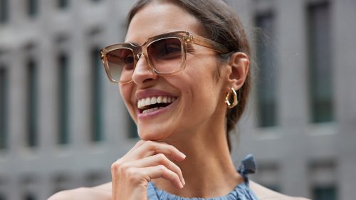happy-young-brunette-european-woman-holds-chin-looks-away-smiles-broadly-being-in-good-mood-wears-trendy-sunglasses-and-fashionable-clothes-poses-against-blurred-background-spends-free-time-in-city-min