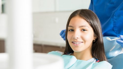 portrait-of-smiling-teenage-girl-with-braces-sitting-on-chair-while-dentist-standing-in-clinic-min